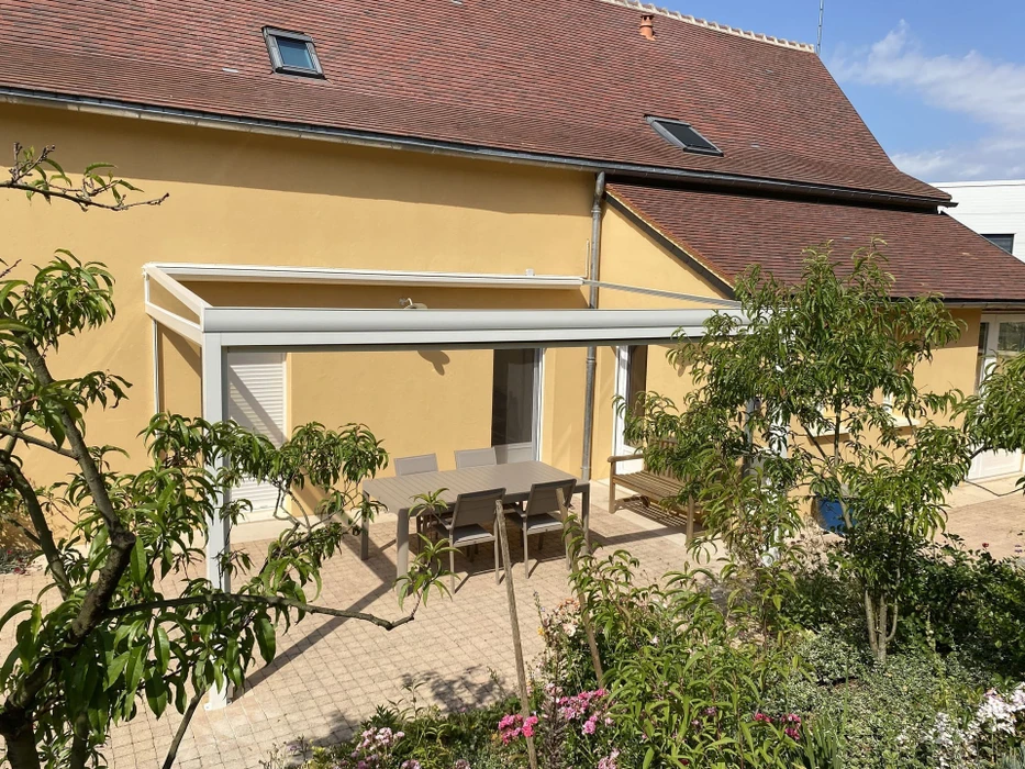 Terrasse extérieure couverte d'une véranda aluminium blanc à toit plat vitré, adossée à une maison enduit jaune ocre à toit en tuiles brunes