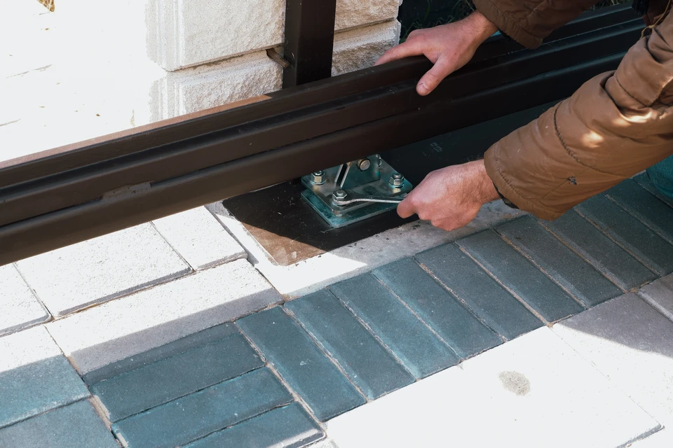 Deux artisans en tenue professionnelle travaillant de concert à l'installation d'un portail battant aluminium sur les piliers d'entrée d'une propriété résidentielle. L'un ajuste un gond tandis que l'autre contrôle l'alignement avec un niveau. Chantier en milieu rural, jour lumineux, montrant le professionnalisme, la précision et la collaboration de l'équipe. L'arrière-plan laisse voir une maison récente avec aménagement paysager soigné.